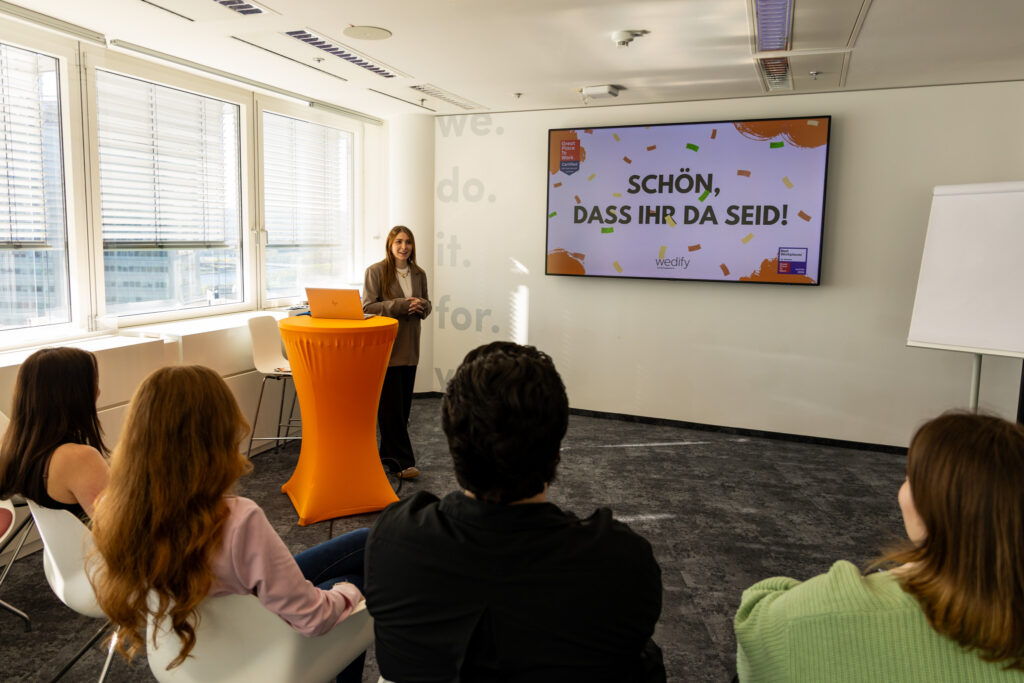 eine junge Frau steht vor einer Gruppe und präsentiert. Auf der Präsentation steht