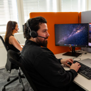 Ein Mitarbeiter sitzt mit Kopfhörern am PC und arbeitet. ER hat schwarze Haaren und einen Bart und trägt ein schwarzes Hemd. Im Hintergrund sieht man eine weitere Mitarbeiterin mit langen braunen Haaren