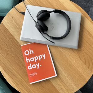 Auf einem Holztisch liegen ein Laptop, ein Headset und ein Notizbuch auf dem steht