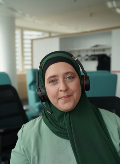 eine Frau mit grünem Kopftuch und türkisem Gewand lächelt in die Kamera, sie trägt ein Headset
