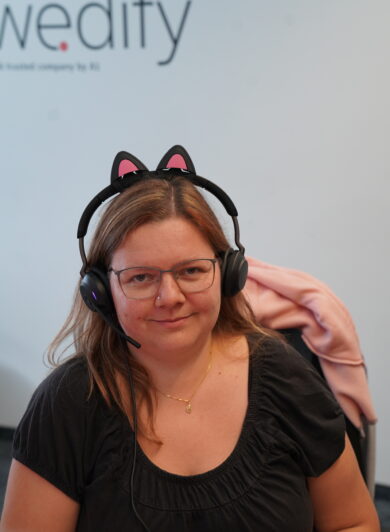 eine Frau mit heller Haut sitzt am Arbeitsplatz und trägt ihr Headset. Auf ihrem Headset sind kleine Katzenohren fixiert, sie lächelt in die Kamera