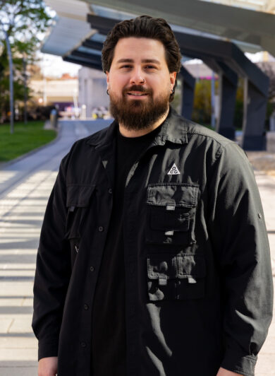 Ein junger Mann mit dunkelbrauenen Haaren und Vollbart im Portrait; er trägt ein schwarzes Hemd