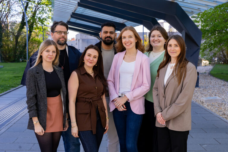 ein Gruppenfoto mit 7 Personen vor urbanem Hintergrund. Es sind 5 Frauen und 2 Männer abgebildet, die alle lächeln