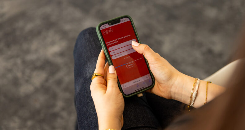 eine Hand mit goldenem Schmuck, die ein Handy hält auf dem eine Umfrage zu sehen ist
