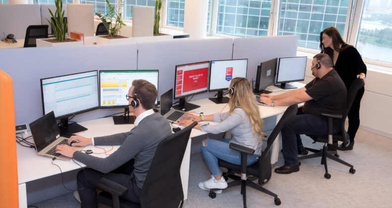 3 Mitarbeiter*innen sitzen vor PC-Bildschirmen und arbeiten. Eine Führungskraft steht unterstützend bei ihnen.