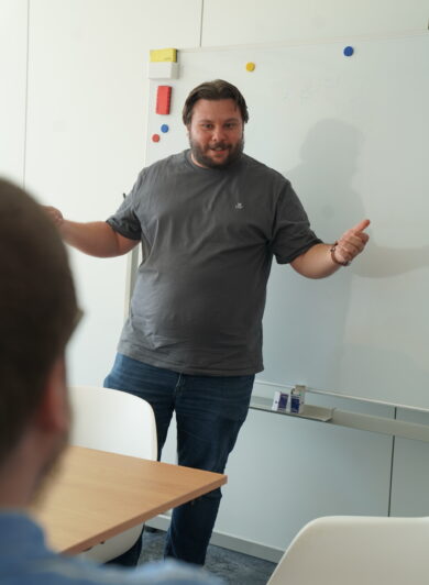 Ein Mann im grauen Tshirt steht vor einem Whiteboard und erklärt einer Person etwas