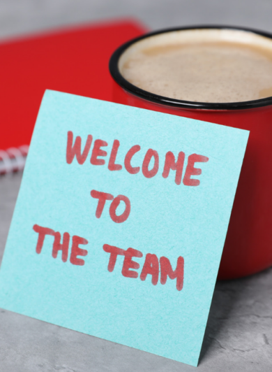 eine Rote Tasse, ein rotes Notizbuch und ein Blaues Schild auf dem steht: Welcome to the team